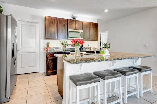 kitchen with stone counters, a kitchen bar, light tile patterned flooring, an island with sink, and stainless steel appliances