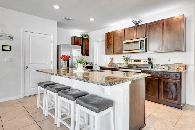 kitchen with light tile patterned floors, stainless steel appliances, a center island with sink, and dark stone counters