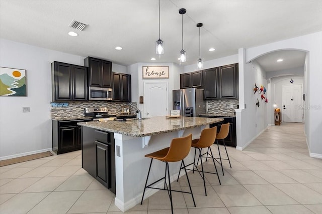 kitchen with light tile patterned floors, appliances with stainless steel finishes, hanging light fixtures, an island with sink, and decorative backsplash