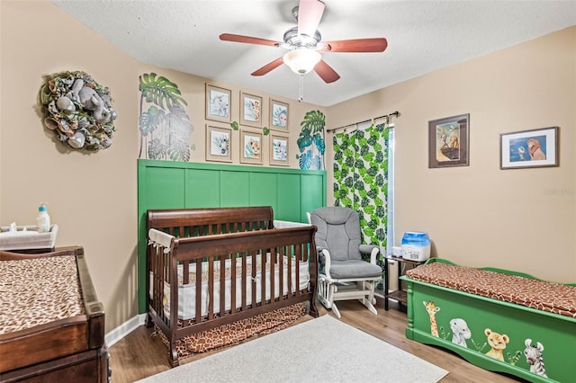 bedroom with ceiling fan, a textured ceiling, a nursery area, and hardwood / wood-style floors