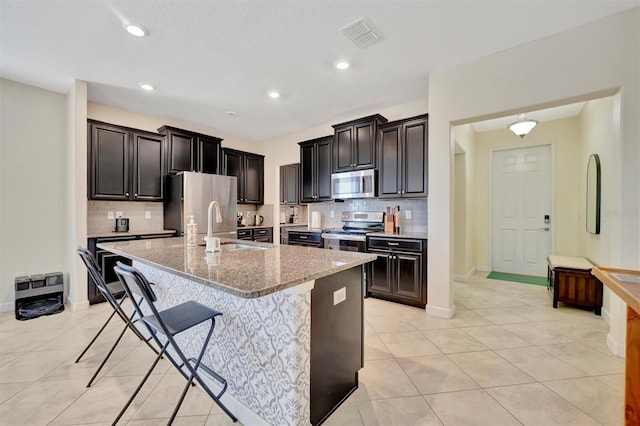 kitchen featuring a kitchen island with sink, light stone countertops, a kitchen breakfast bar, stainless steel appliances, and light tile patterned floors