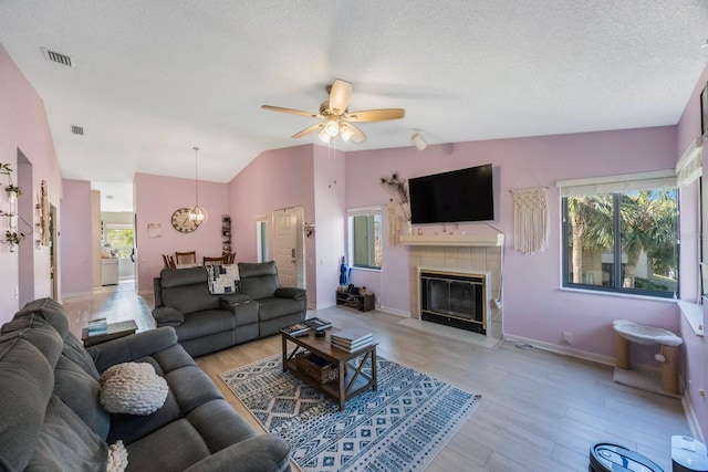 living room featuring vaulted ceiling, light hardwood / wood-style flooring, a tiled fireplace, a textured ceiling, and ceiling fan with notable chandelier