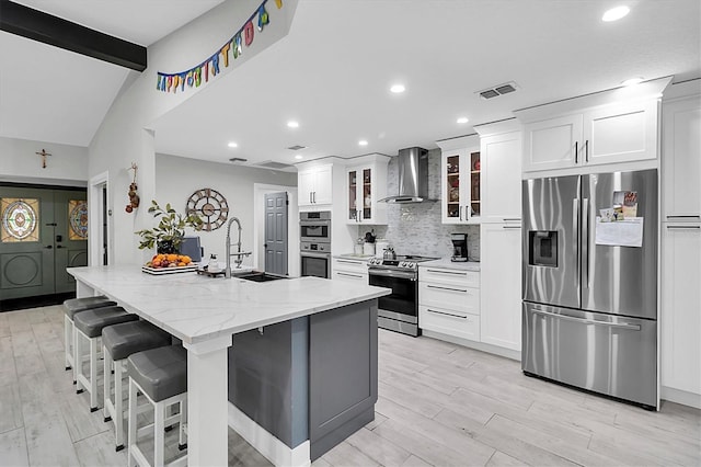 kitchen featuring appliances with stainless steel finishes, wall chimney exhaust hood, beamed ceiling, an island with sink, and sink