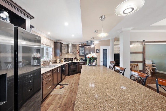 kitchen with a barn door, wall chimney range hood, pendant lighting, black appliances, and sink