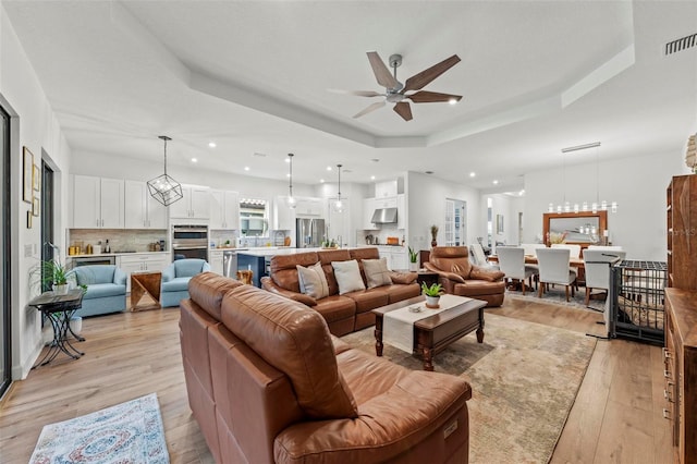 living room with a raised ceiling, ceiling fan with notable chandelier, and light wood-type flooring