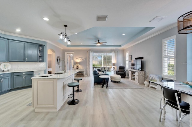 kitchen with light stone countertops, pendant lighting, light hardwood / wood-style flooring, a tray ceiling, and a center island with sink