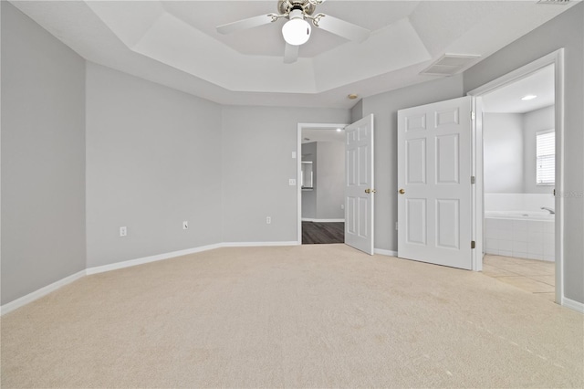 unfurnished bedroom with light carpet, ceiling fan, a tray ceiling, and ensuite bath