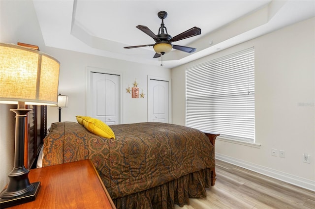 bedroom with a ceiling fan, baseboards, light wood-style floors, a tray ceiling, and two closets