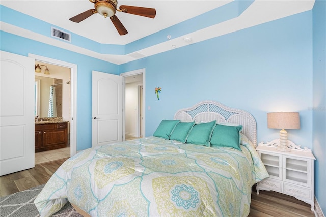 bedroom with ceiling fan, ensuite bath, wood finished floors, visible vents, and a raised ceiling