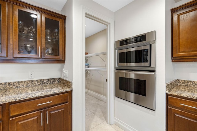 kitchen featuring light stone counters, light tile patterned floors, double oven, brown cabinetry, and glass insert cabinets