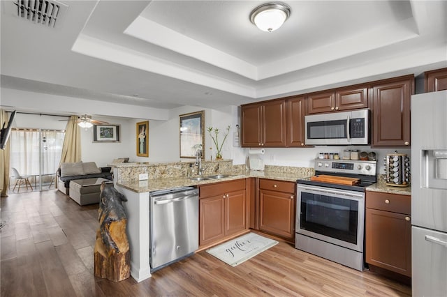 kitchen with kitchen peninsula, appliances with stainless steel finishes, a tray ceiling, light wood-type flooring, and light stone countertops