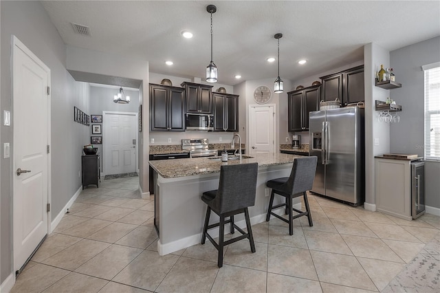 kitchen with an island with sink, hanging light fixtures, light tile patterned floors, stainless steel appliances, and light stone countertops