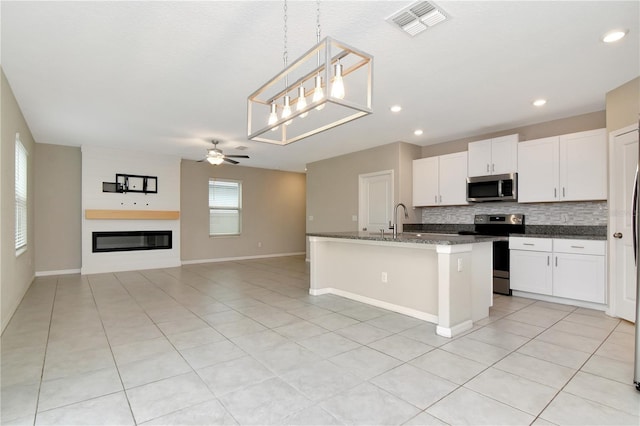 kitchen with stainless steel appliances, hanging light fixtures, a glass covered fireplace, dark stone countertops, and an island with sink