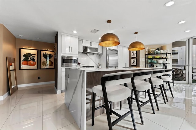 kitchen with pendant lighting, light tile patterned flooring, built in appliances, white cabinetry, and a kitchen island with sink