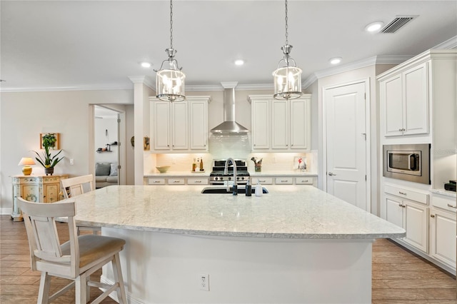 kitchen with stainless steel microwave, decorative light fixtures, white cabinetry, a kitchen island with sink, and wall chimney exhaust hood