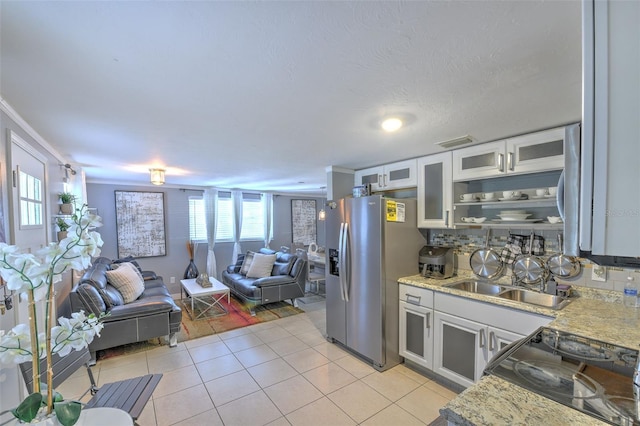 kitchen with stainless steel refrigerator with ice dispenser, glass insert cabinets, open floor plan, light tile patterned flooring, and a sink