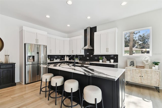 kitchen with appliances with stainless steel finishes, wall chimney range hood, light stone countertops, a kitchen island with sink, and white cabinets