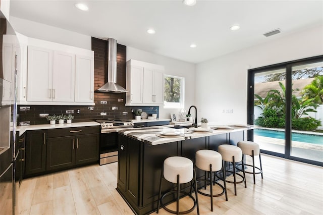 kitchen with sink, stainless steel gas stove, white cabinetry, an island with sink, and wall chimney exhaust hood