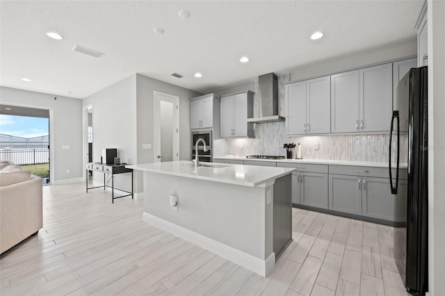 kitchen featuring wall chimney exhaust hood, an island with sink, decorative backsplash, sink, and black fridge