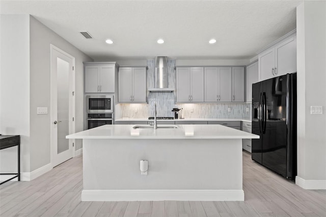 kitchen with black refrigerator with ice dispenser, wall chimney range hood, a center island with sink, and stainless steel oven