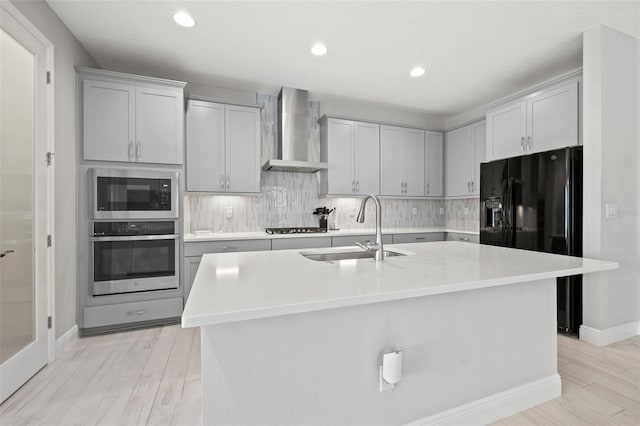 kitchen featuring appliances with stainless steel finishes, sink, gray cabinetry, wall chimney range hood, and a center island with sink