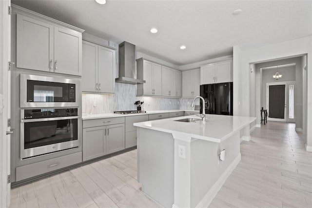 kitchen featuring wall chimney range hood, sink, stainless steel appliances, a center island with sink, and gray cabinets