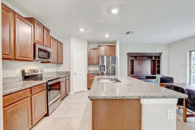 kitchen featuring light tile patterned flooring, an island with sink, sink, stainless steel appliances, and light stone countertops