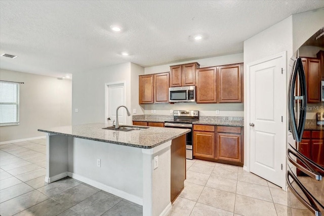 kitchen with sink, a kitchen island with sink, light tile patterned floors, stainless steel appliances, and light stone countertops