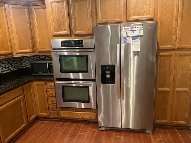 kitchen featuring dark wood-style flooring, backsplash, appliances with stainless steel finishes, brown cabinetry, and dark stone counters