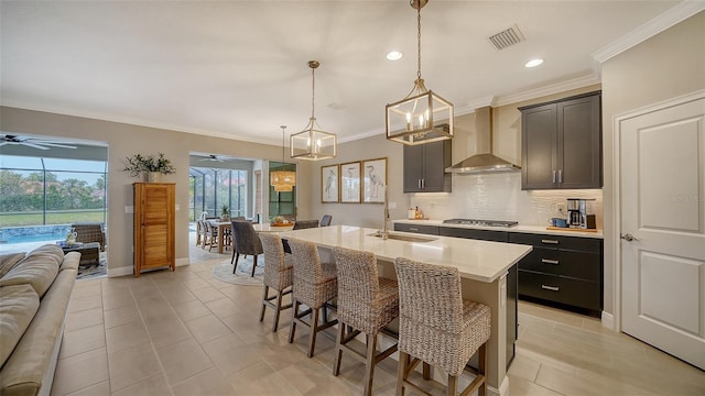 kitchen featuring wall chimney exhaust hood, a kitchen bar, sink, hanging light fixtures, and a kitchen island with sink