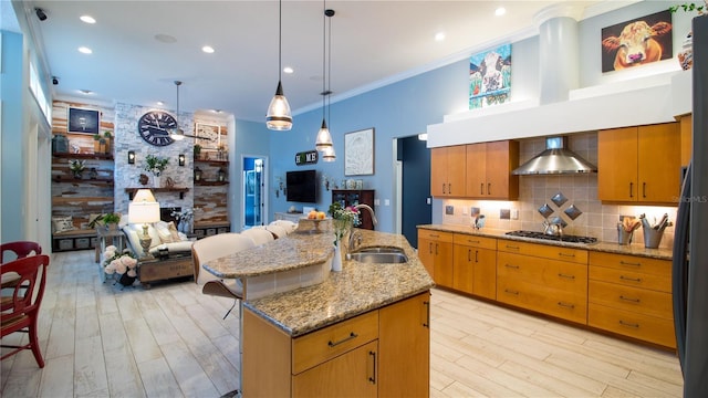 kitchen with wall chimney range hood, sink, a kitchen island with sink, a stone fireplace, and stainless steel gas stovetop