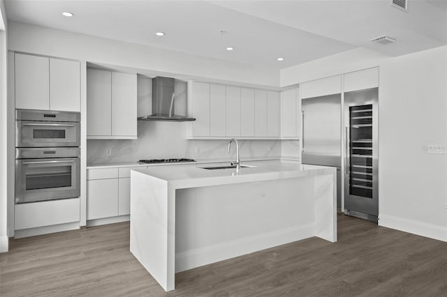 kitchen featuring wall chimney range hood, sink, white cabinetry, hardwood / wood-style flooring, and an island with sink