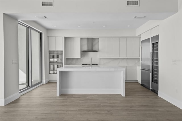 kitchen featuring white cabinetry, an island with sink, appliances with stainless steel finishes, backsplash, and wall chimney exhaust hood