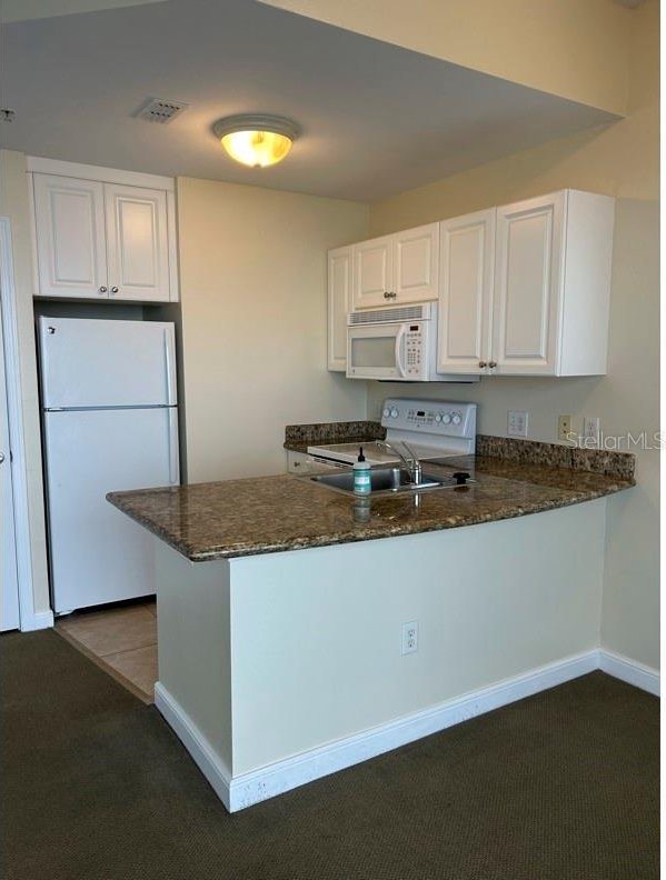 kitchen with dark colored carpet, dark stone countertops, kitchen peninsula, white appliances, and white cabinets