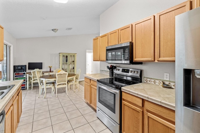 kitchen with stainless steel appliances, tile countertops, light brown cabinets, and light tile patterned floors