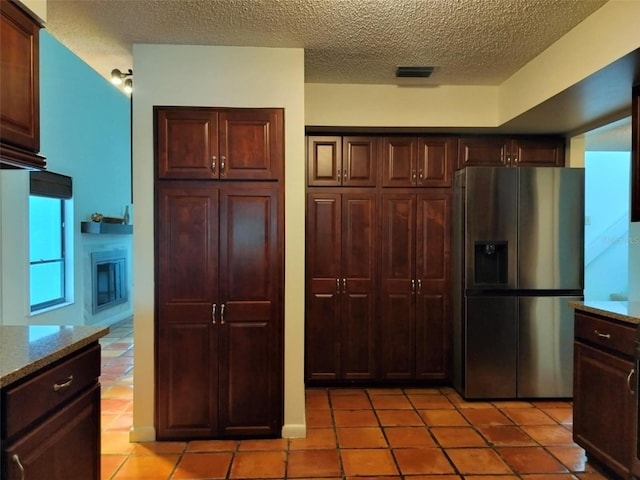 kitchen with stainless steel fridge, light tile patterned flooring, dark brown cabinetry, a textured ceiling, and light stone countertops