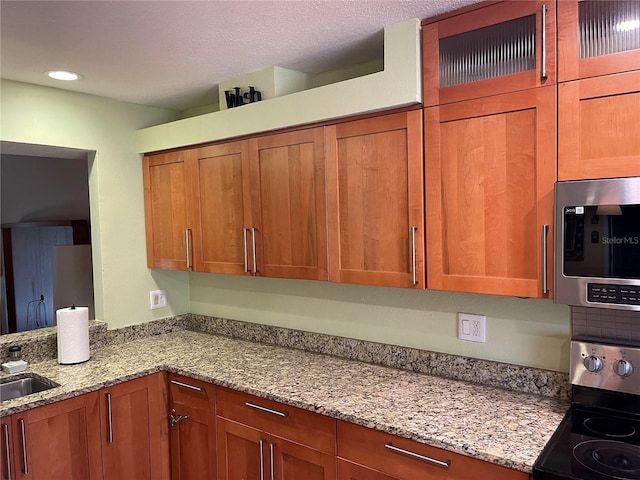 kitchen with stainless steel appliances, brown cabinetry, glass insert cabinets, a sink, and light stone countertops