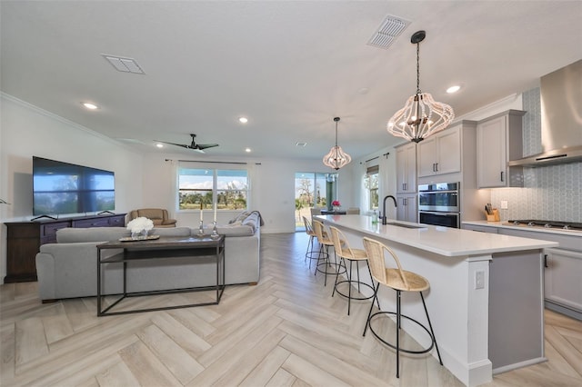 kitchen featuring wall chimney range hood, gray cabinetry, hanging light fixtures, a large island with sink, and light parquet flooring