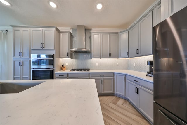 kitchen with wall chimney exhaust hood, gray cabinetry, light stone counters, tasteful backsplash, and appliances with stainless steel finishes