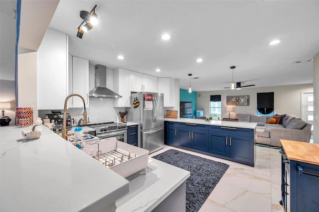 kitchen featuring blue cabinets, tasteful backsplash, kitchen peninsula, stainless steel appliances, and wall chimney range hood