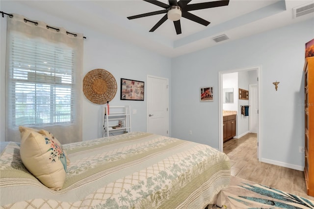 bedroom featuring ceiling fan, ensuite bathroom, a tray ceiling, and light wood-type flooring