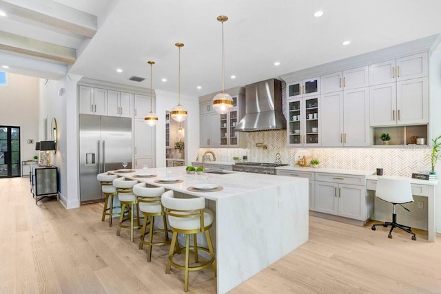 kitchen with sink, hanging light fixtures, appliances with stainless steel finishes, a kitchen island with sink, and wall chimney range hood