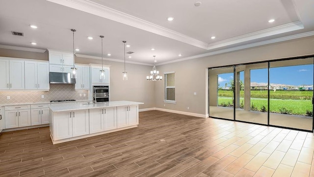 kitchen with decorative light fixtures, white cabinets, a kitchen island with sink, gas stovetop, and range hood