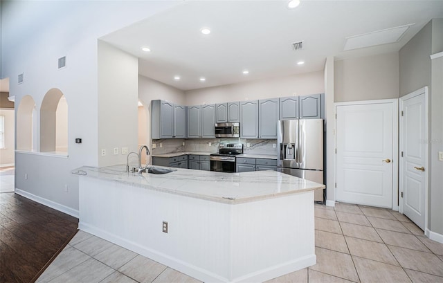 kitchen with gray cabinets, appliances with stainless steel finishes, sink, kitchen peninsula, and light stone countertops