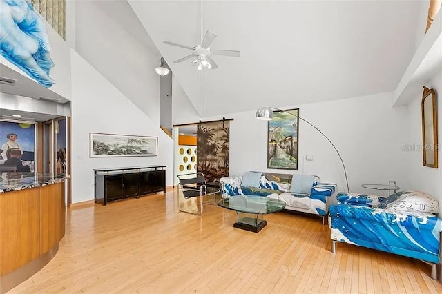 living room with ceiling fan, a barn door, high vaulted ceiling, and light wood-type flooring