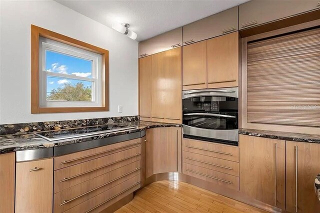 kitchen featuring black electric cooktop, stainless steel oven, dark stone counters, and light wood-type flooring