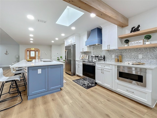 kitchen with appliances with stainless steel finishes, beamed ceiling, white cabinets, custom exhaust hood, and a kitchen island with sink