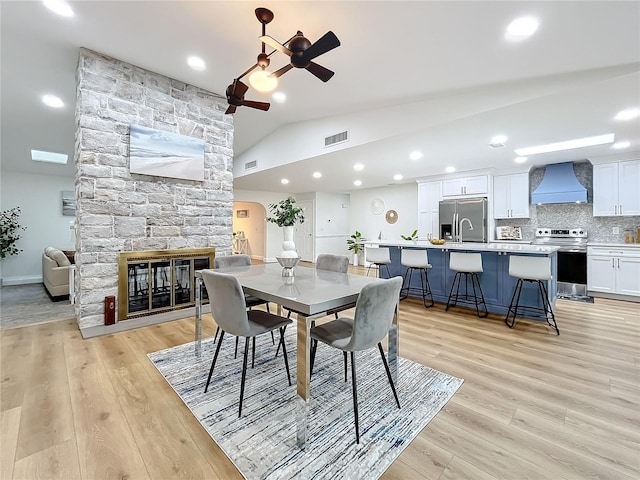 dining space with ceiling fan, a stone fireplace, light hardwood / wood-style floors, and vaulted ceiling