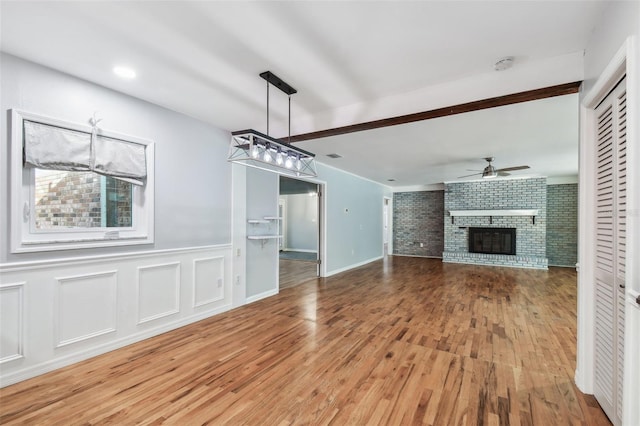 unfurnished living room with a ceiling fan, a brick fireplace, wainscoting, and light wood-style flooring