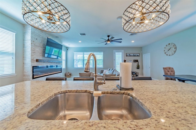 kitchen with sink, ceiling fan with notable chandelier, a stone fireplace, and light stone counters